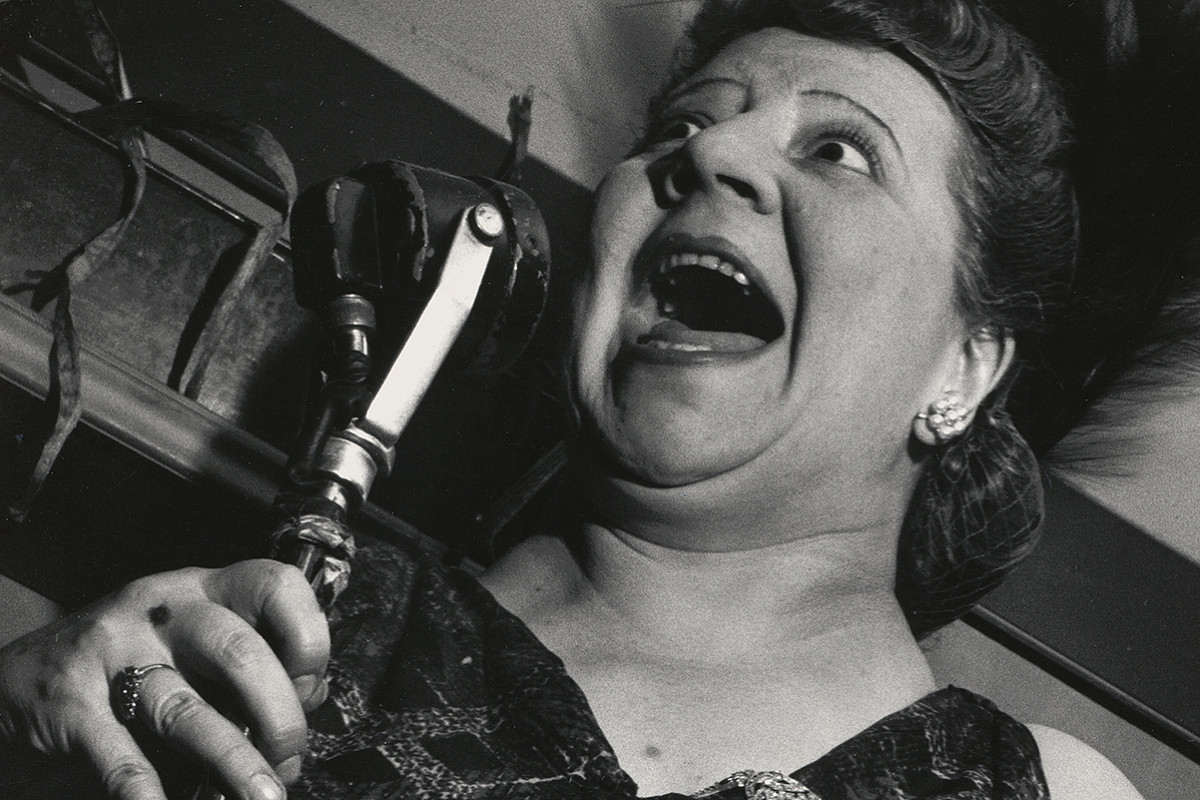 A woman in an evening dress holding a standing microphone up to her face. Her mouth is wide open.