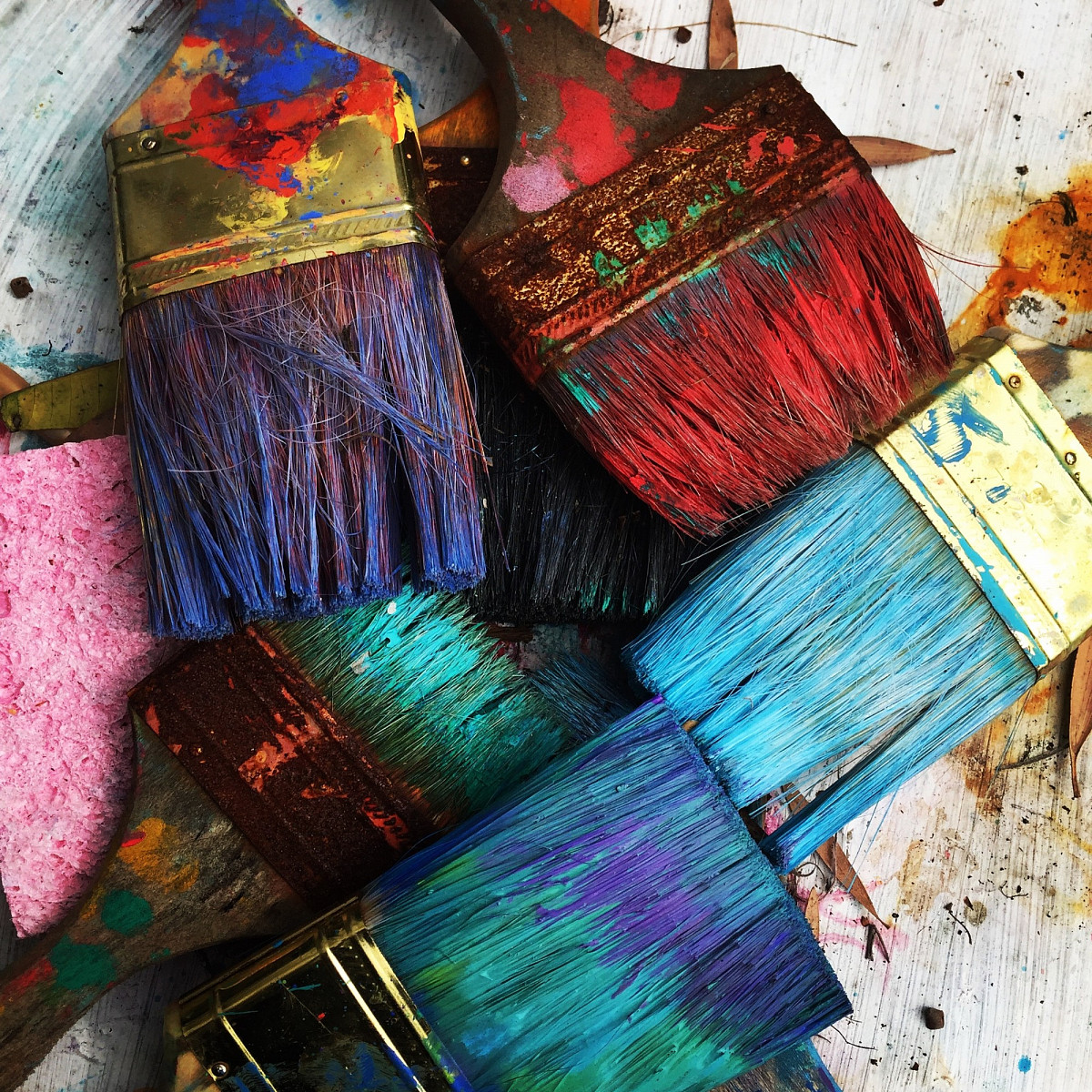Several wide brushes, each soaked in a different color. Underneath a wooden floor and a pink sponge | Unsplash © Rhondak-Unsplash.org