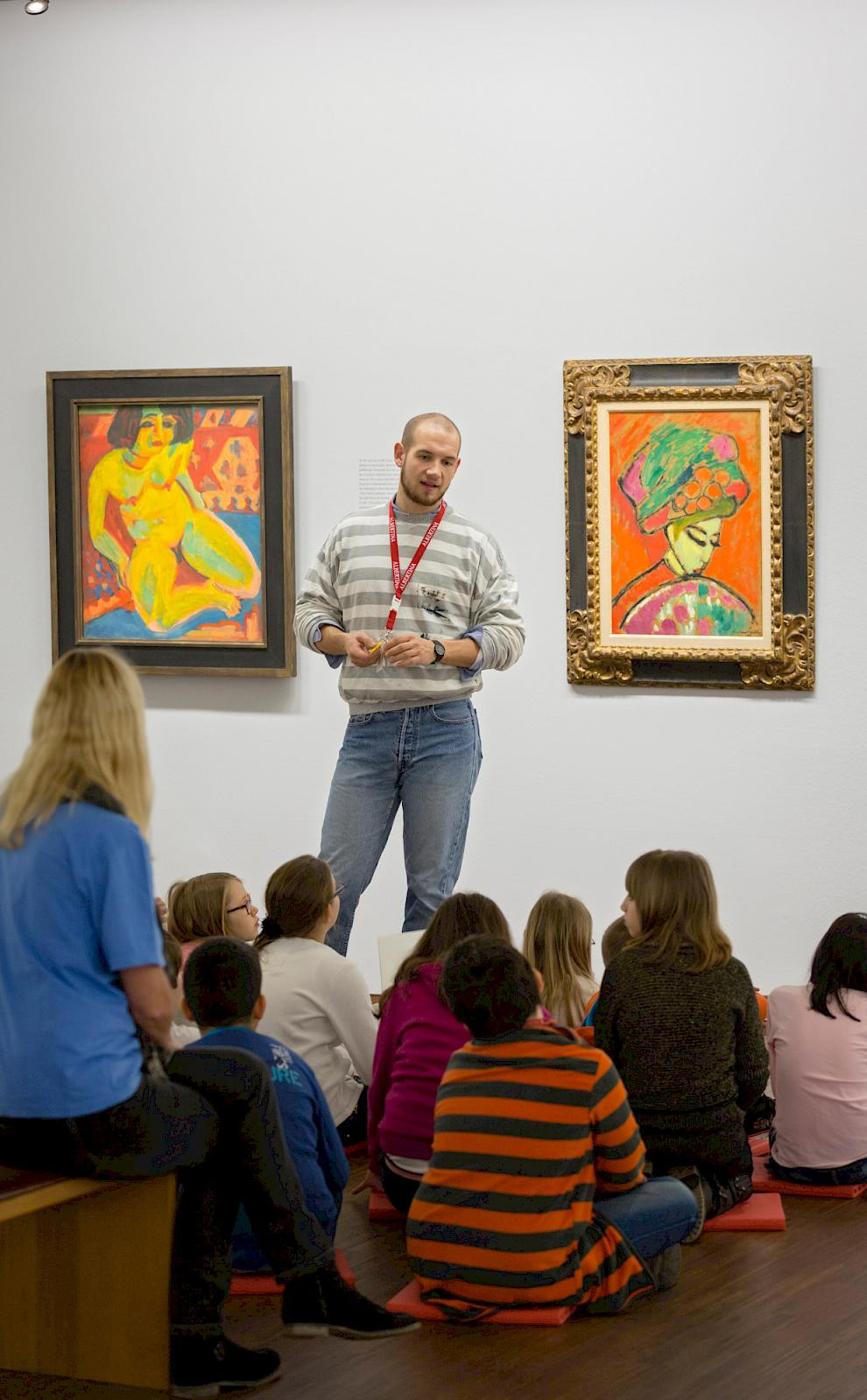 An art educator stands in front of a seated group of children in a museum room and explains two colorful paintings on the wall. © Rainer Mirau