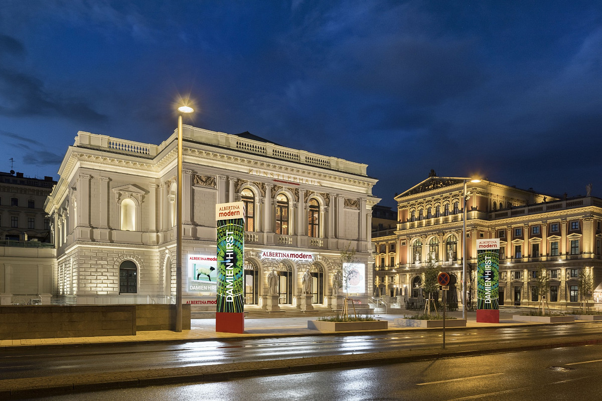 Mockup der ALBERTINA MODERN bei Nacht. Dunkler Himmel, beleuchtetes Gebäude. Die aktuellen Ausstellungen sind in den erhellten Säulen präsentiert..