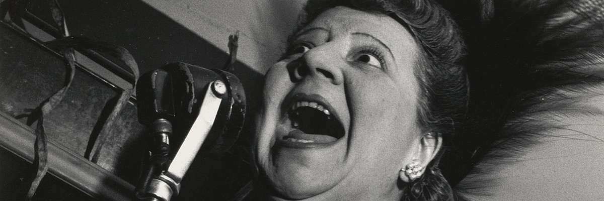 Eine Frau im Abendkleid, die ein Stehmikrofon zum Gesicht hält. Ihr Mund ist dabei weit geöffnet. Credit: Lisette Model | Sängerin im Cafe Metropole, New York City, 1946 | ALBERTINA Wien © 2025 Estate of Lisette Model, courtesy Lebon, Paris / Keitelman, Brussels