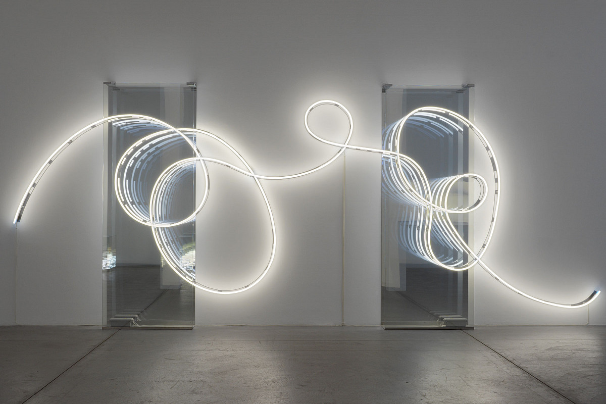 Blick in dunklen Ausstellungsraum, der nur von einer geschwungenen Lichtinstallation vor einem Spiegel beleuchtet wird. | Brigitte Kowanz | Email 02.08.1984 03.08.1984, 2021 | ESTATE BRIGITTE KOWANZ © Estate Brigitte Kowanz / Bildrecht, Wien 2025 | Foto: Peter Hoiss