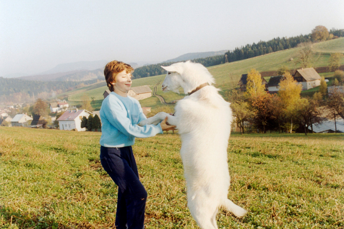Eine Frau steht auf einem Feld. Ihr gegenüber steht eine weiße Ziege aufrecht. Die Frau hält die Vorderbeine des Tieres mit ihren Händen. Credit: Jitka Hanzlová | Untitled, 1993 / aus der Serie: Rokytník, 1990 – 1994 | Courtesy of the artist © Jitka Hanzlová / Bildrecht, Wien 2025