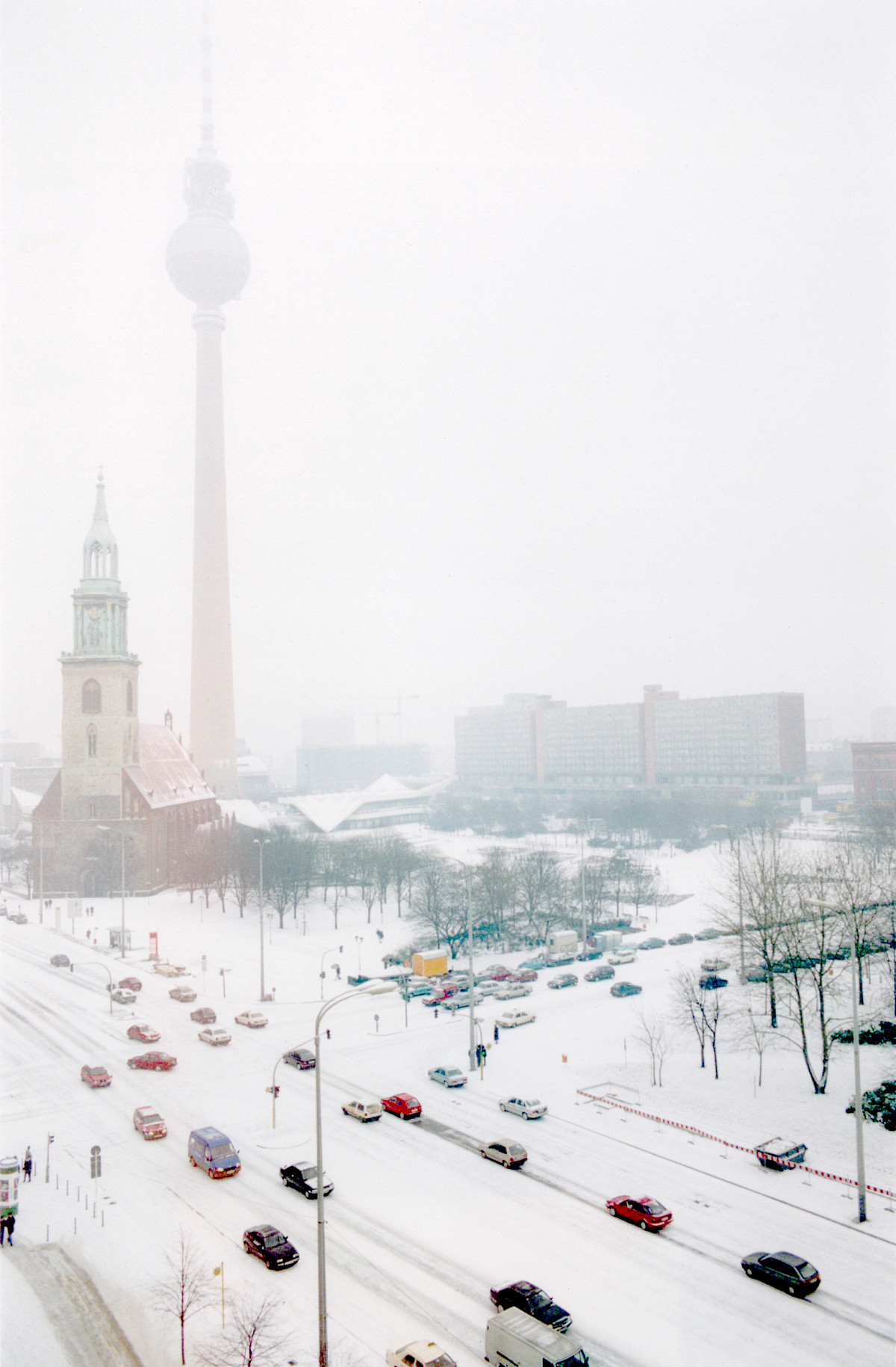 Eine schneebedeckte Straße mit dichtem Autoverkehr. Im Hintergrund eine Kirche, ein Fernsehturm und Blockbauten im Schnee.