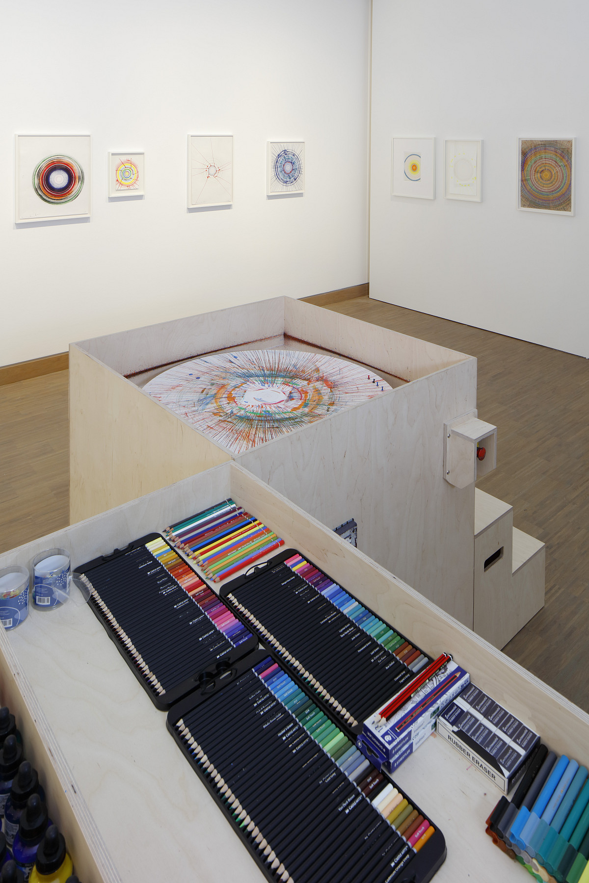 Foto der Zeichenmaschine von Damien Hirst mit einer großen Auswahl an bunten Stiften in einem hellen Ausstellungsraum mit Hirst's ''Spin Drawings'' an der Wand.
