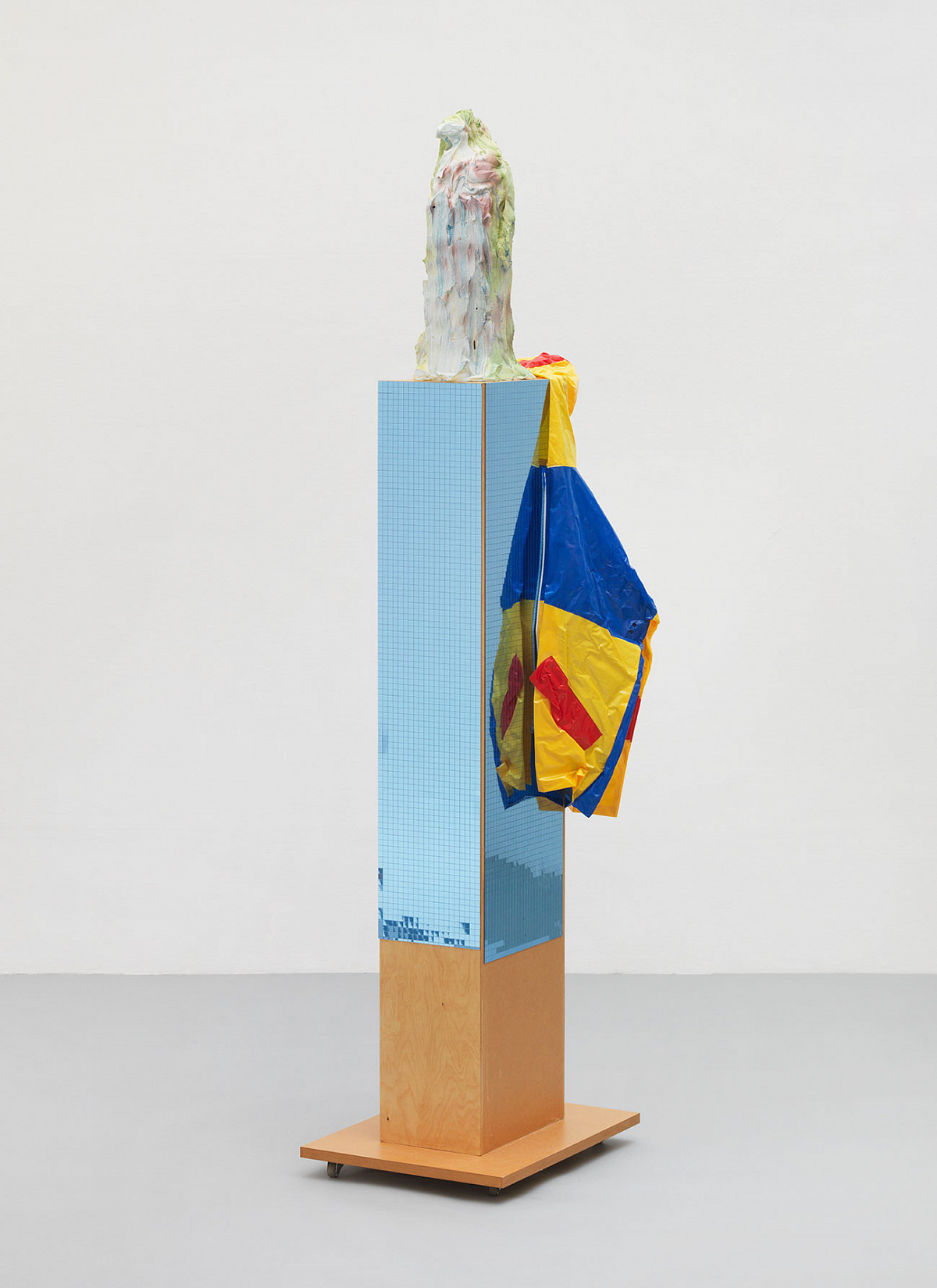 Eine Säule mit Glitzerstreifen, einer Jacke in blau-rot-gelb an einer Kante aufgehängt und einer zusätzlichen, weiß eingefärbten Form auf der Spitze der Säule.
