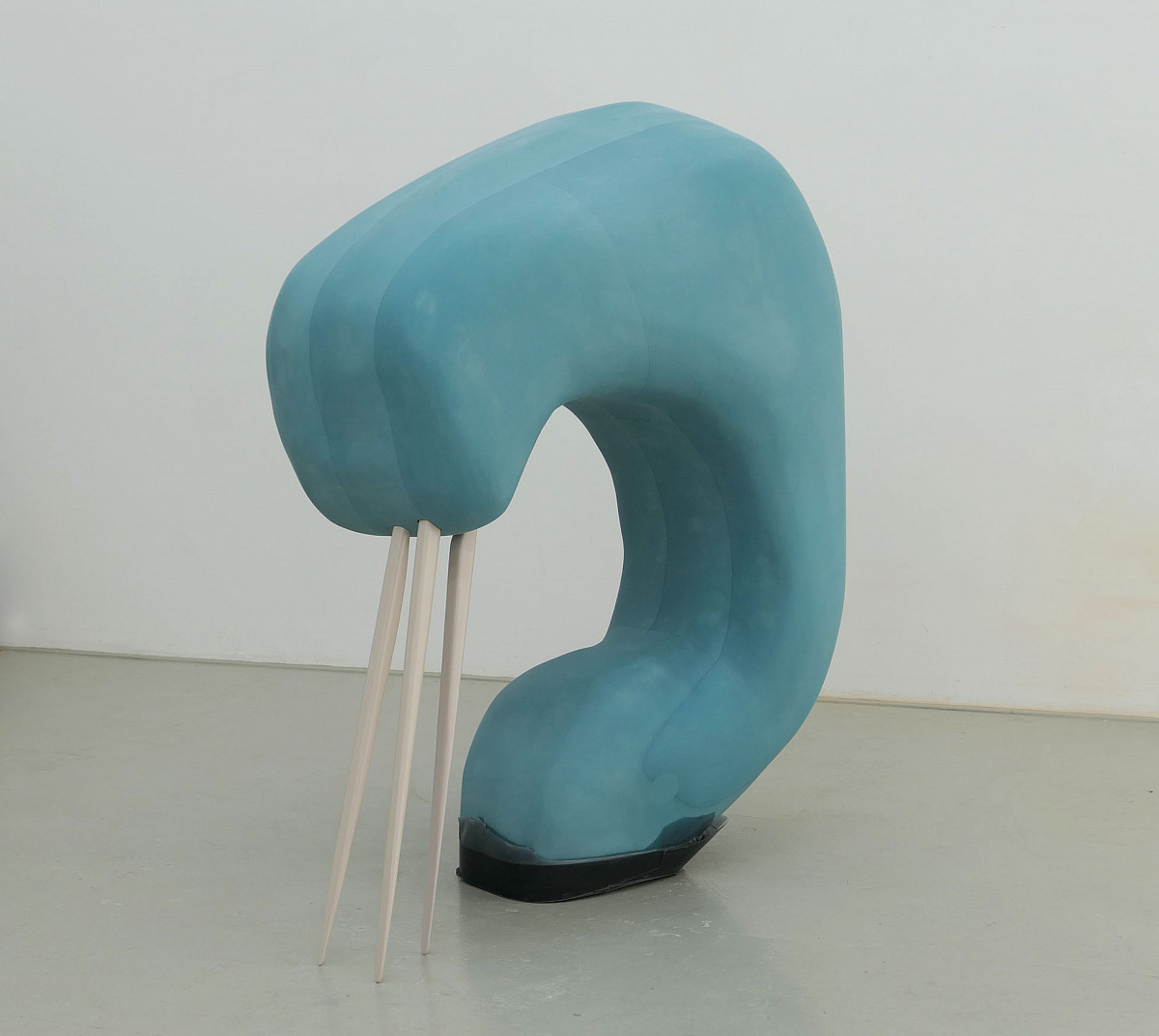 A blue, thick sculpture with a semi-circular shape. Wooden sticks are attached to the upper end, which reach down to the ground and have a pointed end, similar to toothpicks.