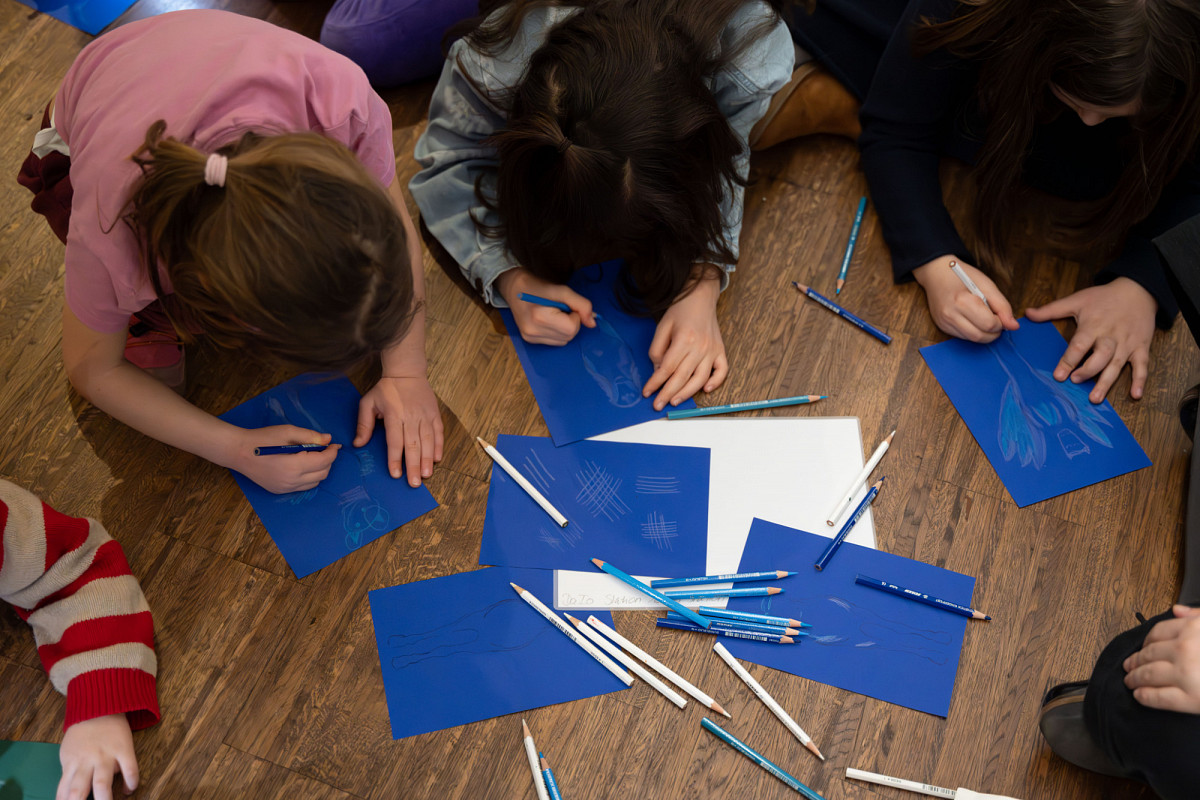 Aus der Vogelperspektive: Kinder sitzen am Boden und malen, vor ihnen liegt blaues und weißes Papier, sowie blaue und weiße Buntstifte