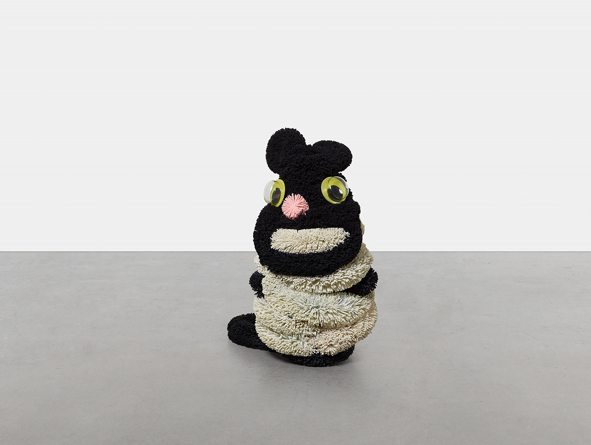 Ein Stofftier, schwarz-weiß mit breitem weißen Mund, zwei Kulleraugen und einer rosanen Nase sitzend in der Ausstellungshalle am grauen Boden mit grauer Wand im Hintergrund.