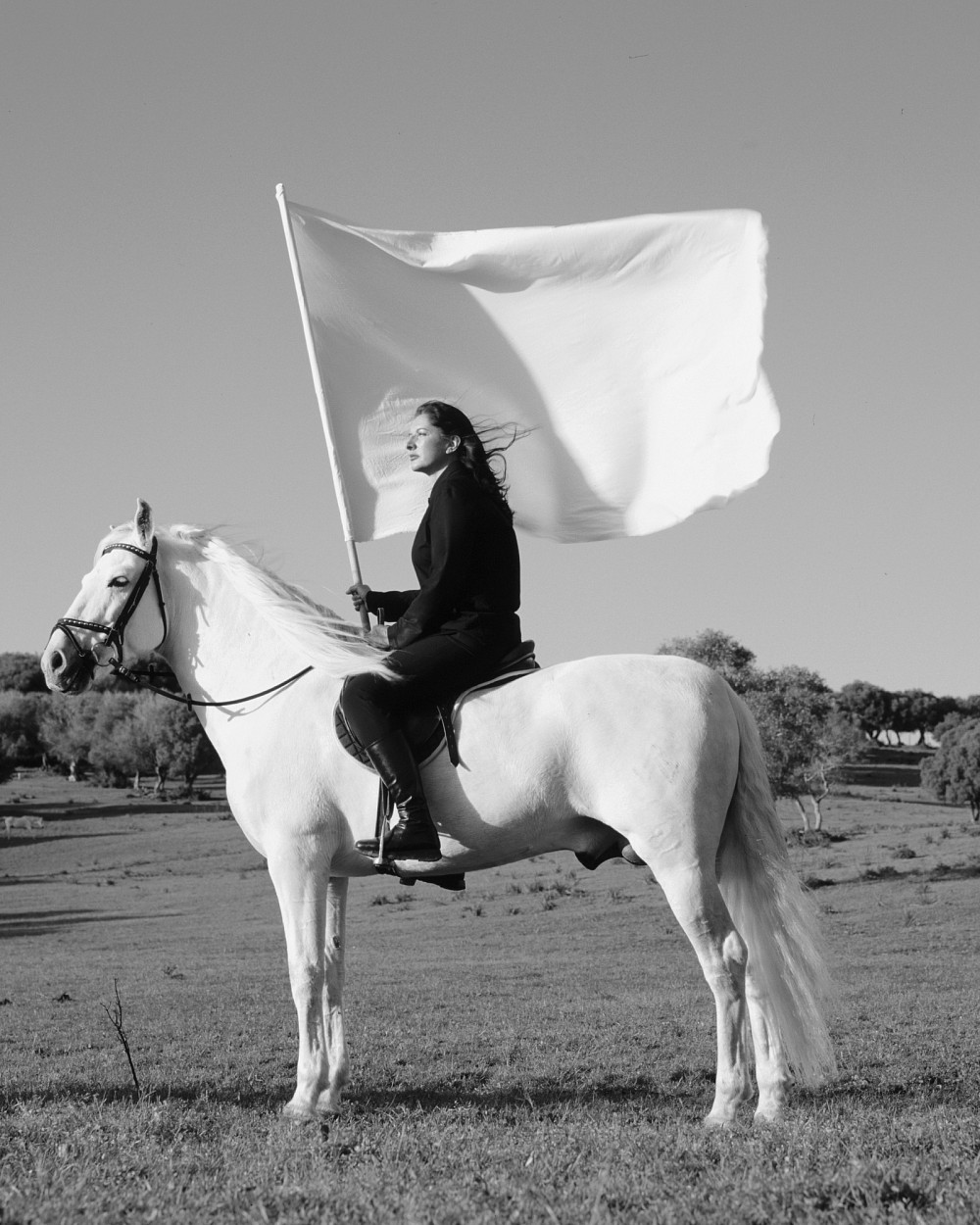 Schwarz-Weiß-Fotografie, auf der die Künstlerin auf einem weißen Pferd sitzt und eine große weiße Fahne hält. | Marina Abramović | The Hero, 2001, Courtesy of the Marina Abramović Archives, and Galeria Luciana Brito © Marina Abramović. Image courtesy of Marina Abramović Archives