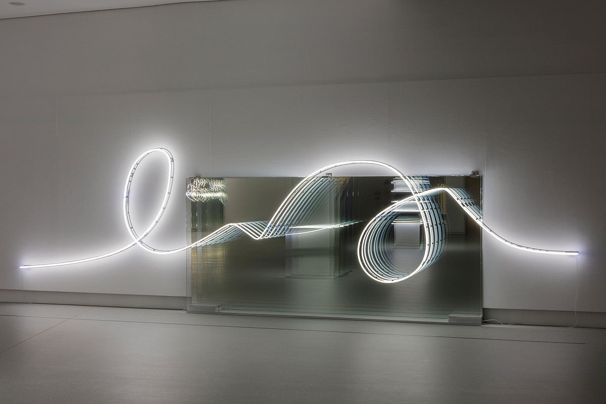 View into a dark exhibition room, illuminated only by a curved light installation in front of a mirror.