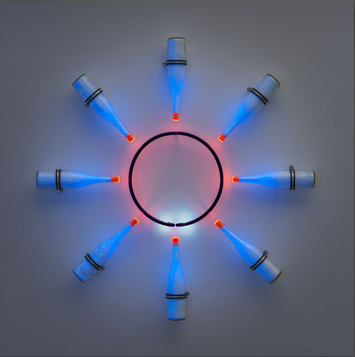 Installation eines schwarzen Kreises, aus dem rotes Licht auf acht um ihn herum platzierte Glasflaschen scheint. Diese scheinen in einem blauen Ton.