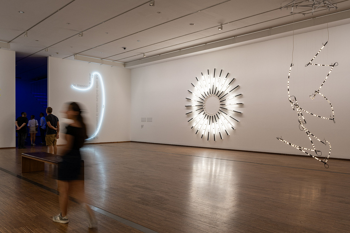 In the hall: Three installations by the artist hang on the wall. A large circle formed from small, straight neon tubes, to the right of it a chain of tubes hanging from the ceiling in different directions. To the left of it, a brightly shining neon tube that winds vertically upward on the wall.