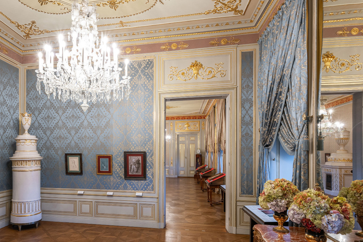 A glimpse into a stately room with blue wallpaper and crystal chandeliers on the ceiling, two large windows with blue curtains, and parquet flooring.