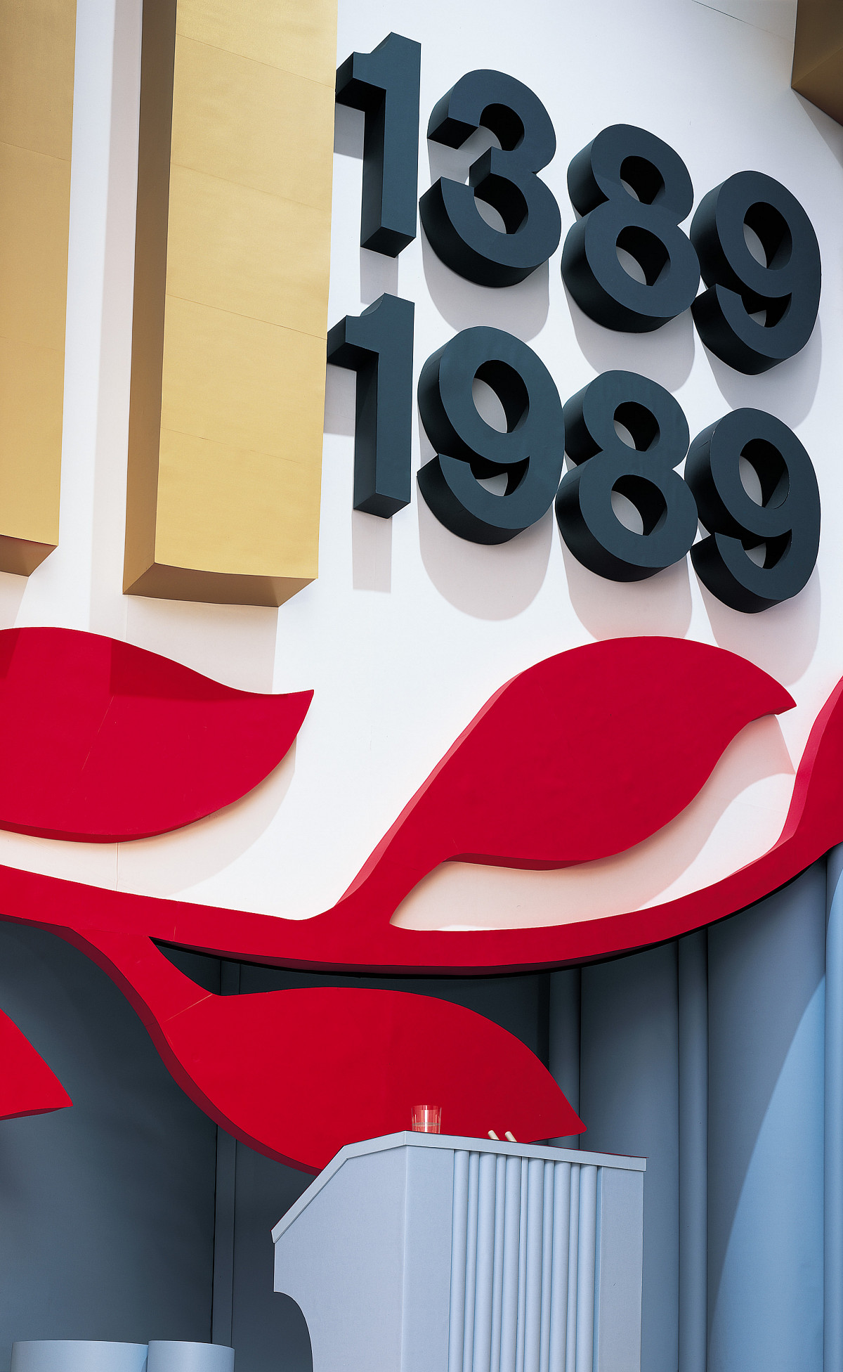 Bild eine Podiumsgestaltung mit Jahreszahlen in schwarz auf weißem Hintergrund, der von roten Blätterranken umgeben ist.