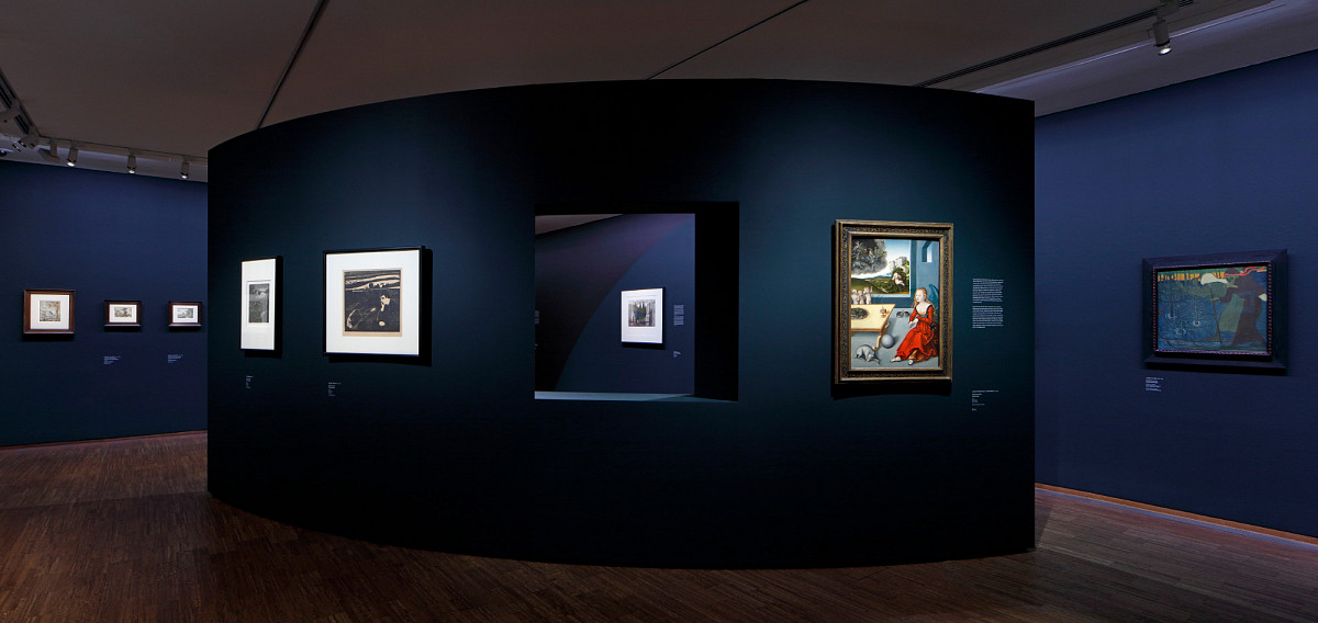 Ein Ausstellungsraum mit dunkelblauer Wandfarbe und in der Mitte eine leicht gewölbte Wand mit einem Fenster, durch das man auf ein Gemälde an der Wand dahinter blicken kann.