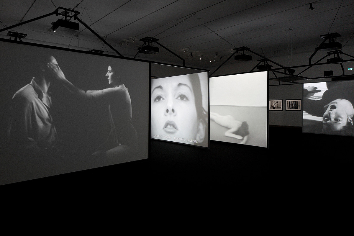 Ansicht der Ausstellungsinstallation: Mehrere Videoleinwände hintereinander aufgehängt. Auf den Leinwänden sieht man schwarz-weiß-Videos, die Aktionen der Künstlerin zeigen.