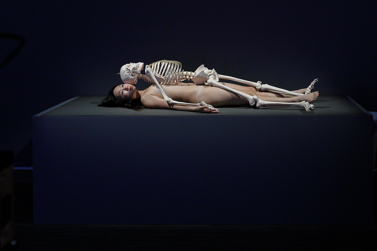 A woman lies naked on a black pedestal. A human skeleton lies on top of her. Both are looking up.