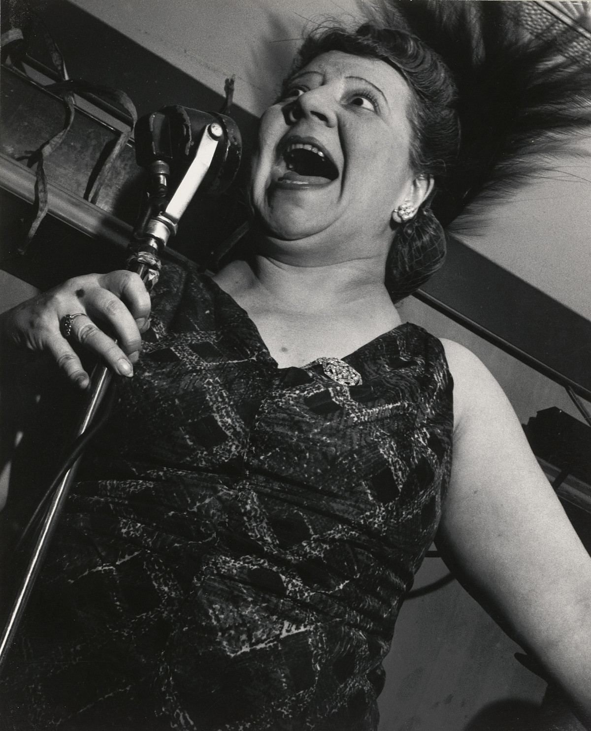 A woman in an evening dress holding a standing microphone up to her face. Her mouth is wide open.