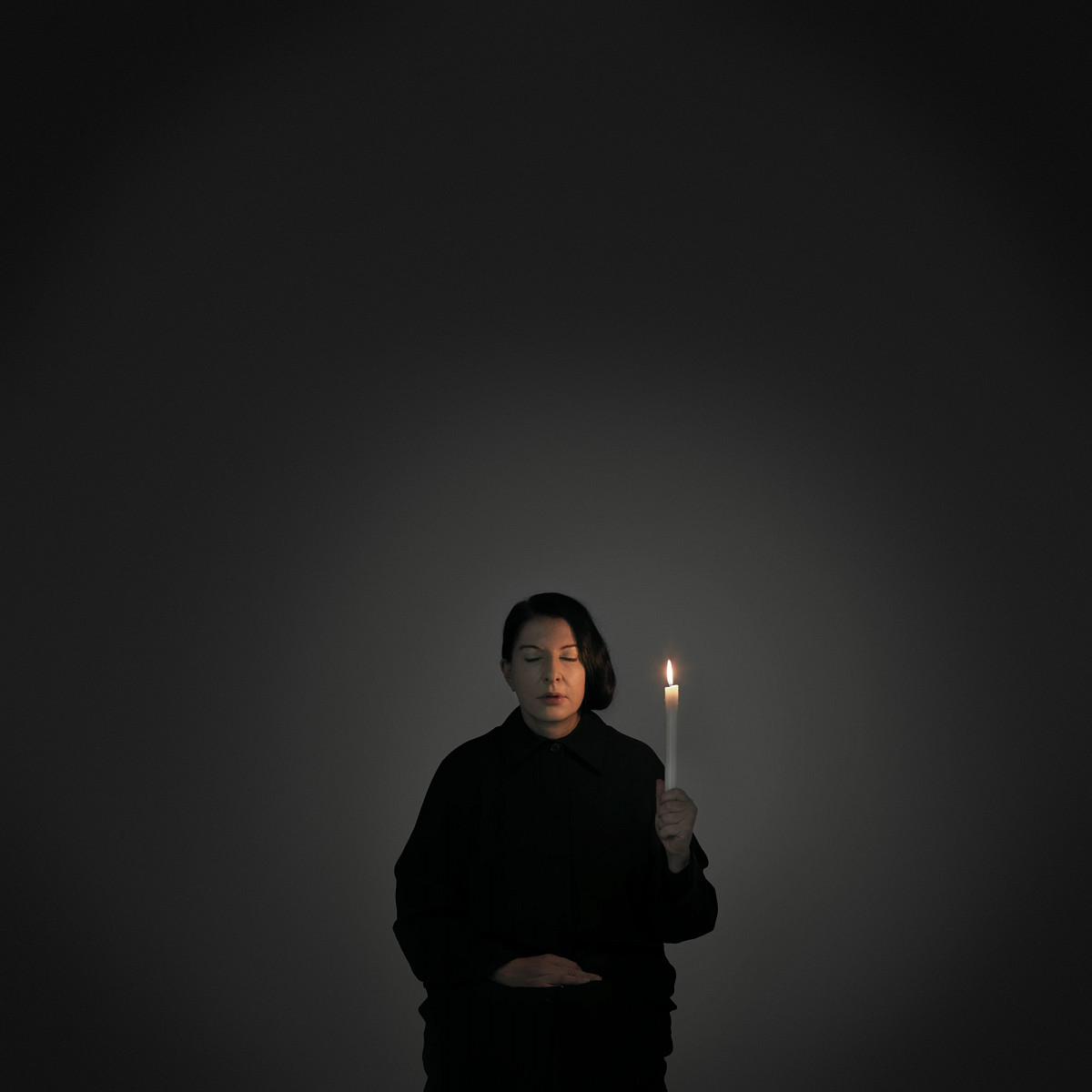 Marina Abramović sitzt schwarz angezogen und mit geschlossenen Augen in einem dunklen Raum, der nur von der Kerze in ihrer Hand erhellt wird. | Credit:Marina Abramović | Artist Portrait with a Candle (A), 2012 | Courtesy of the Marina Abramović Archives, and Galerie Krinzinger | © Courtesy of the Marina Abramović Archives / Bildrecht, Wien 2025