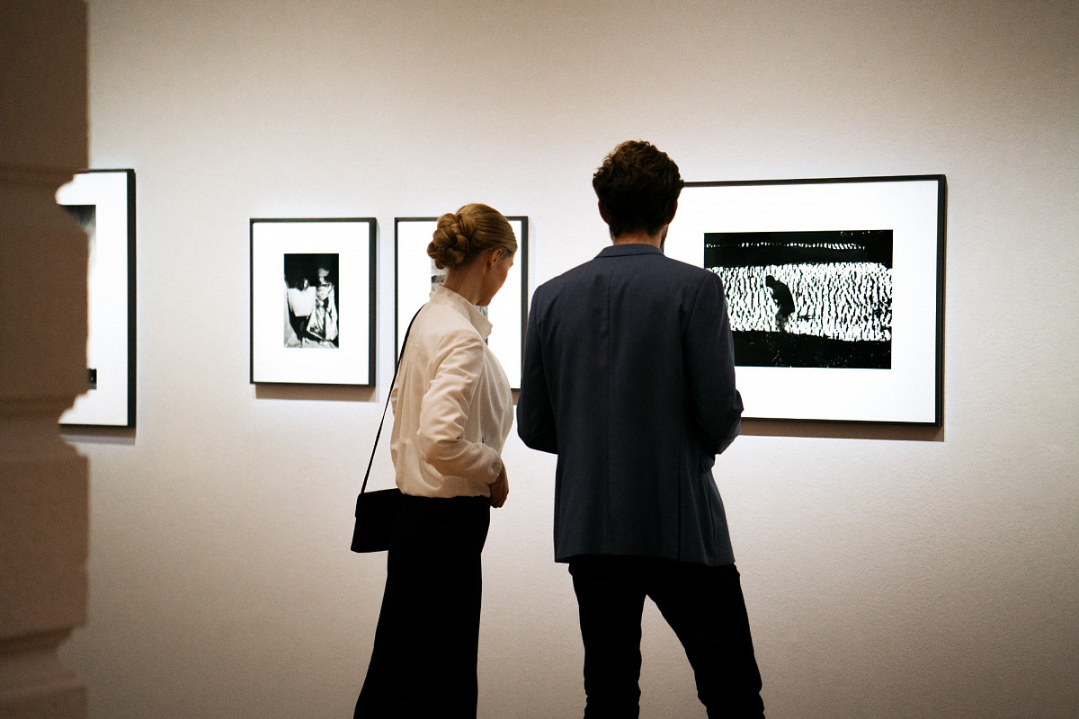 Zwei Personen in der Ausstellungshalle, vor ihnen drei schwarz-weiße Bilder in schwarzem Rahmen | Foto © Marko Mestrovic