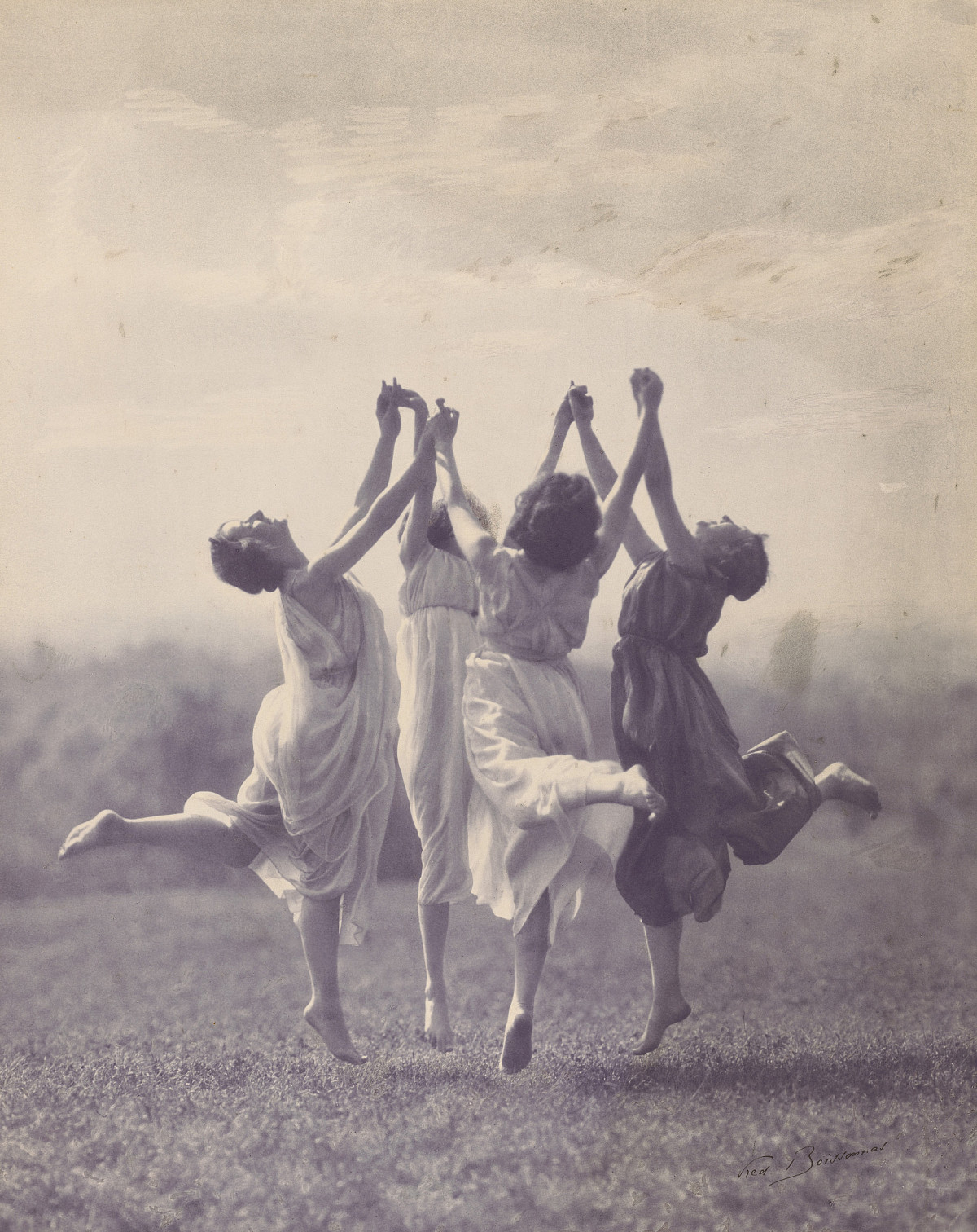 Photograph of four women in flowing dresses jumping on a meadow. They each stretch out one leg and hold hands. They stretch their hands toward the sky.