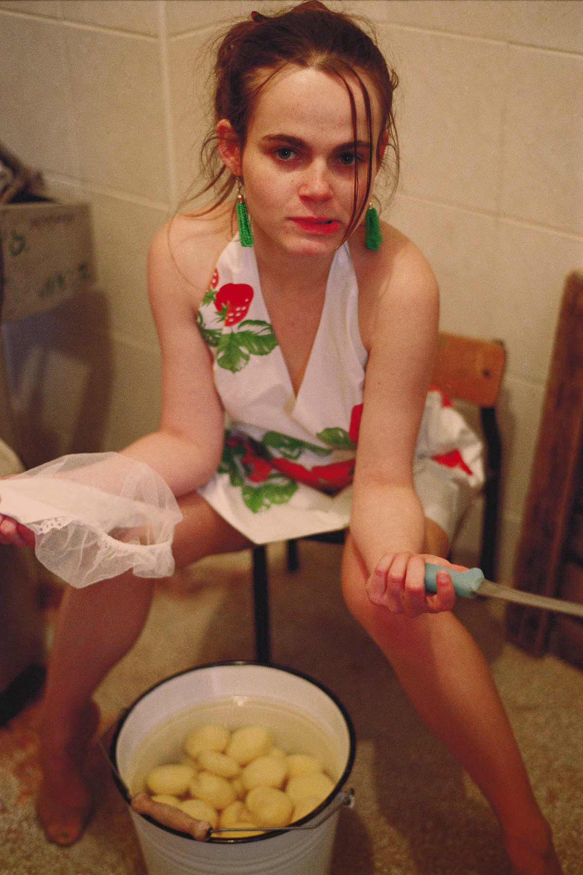 Eine junge Frau auf einem Stuhl sitzend mit geöffneten Beinen. Unter ihr steht ein Kübel mit geschälten Kartoffeln. Sie hält ein Messer und ein nasses Tuch in der Hand.