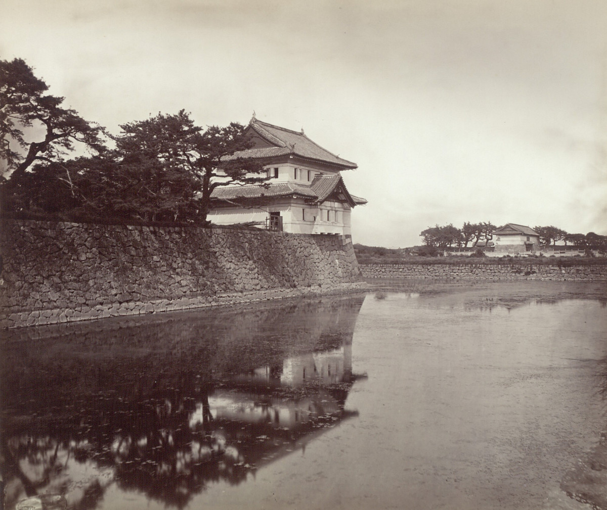 Black-and-white photograph of a Japanese-style guardhouse on the edge of the moat, surrounded by trees.