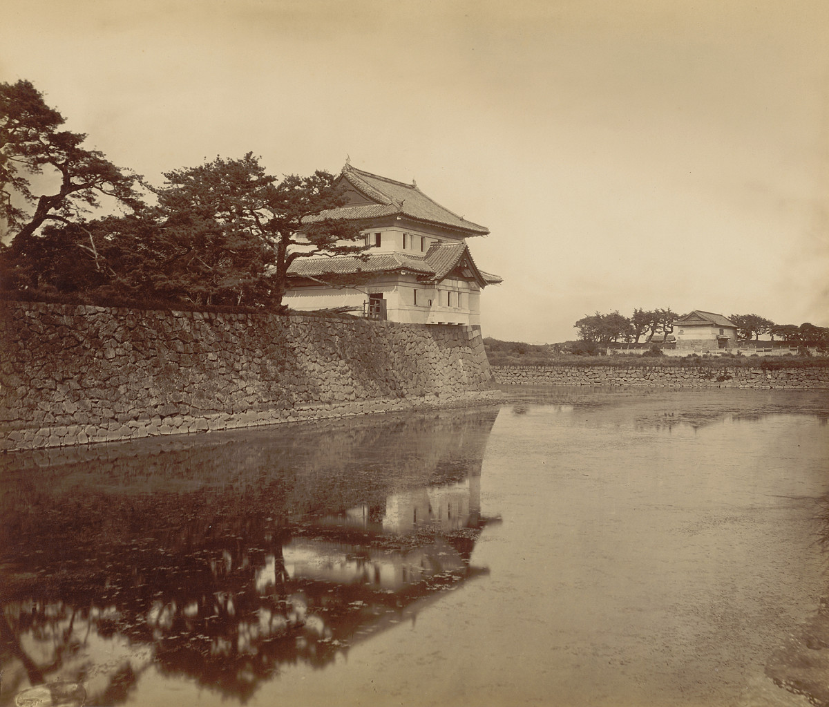 Schwarz-Weiß-Fotografie eines Wachgebäudes im japanischen Stil am Rande des Burggrabens und von Bäumen umgeben.