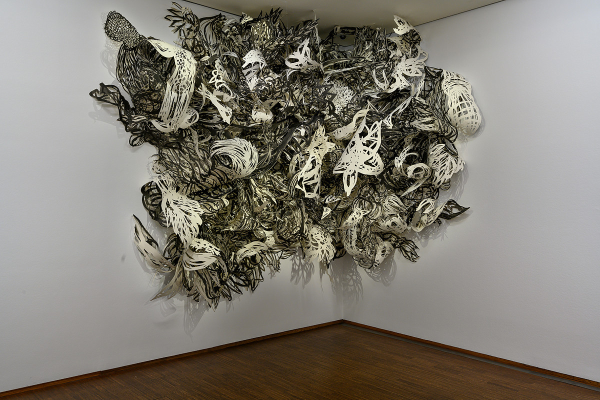 Eine Ecke eines Raumes mit braunem Parkettboden und weißer Wand. An der Wand, in der Ecke, hängt eine großflächige Installation aus Papierschnitten in verschiedenen Mustern in Schwar-Weiß.