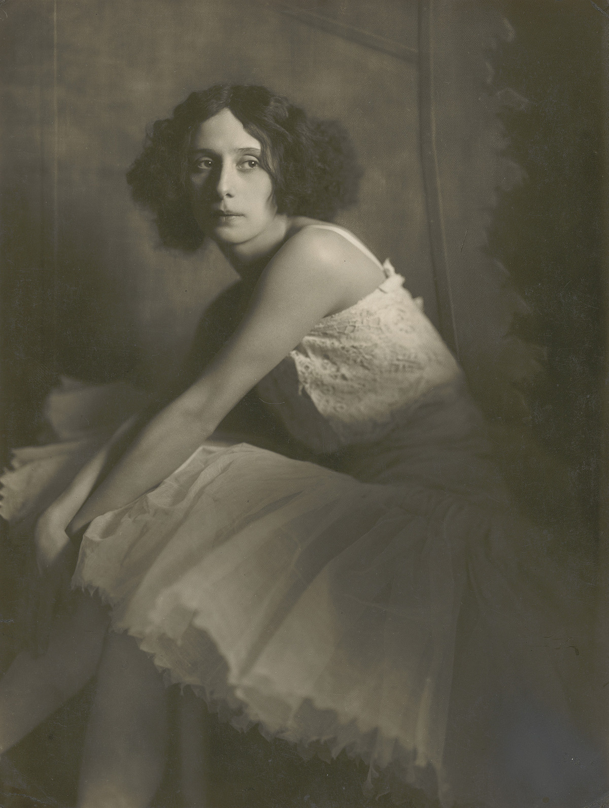 A woman with chin-length, wavy hair wearing a white strapless dress with a flared skirt, sitting with her hands stretched out in her lap and looking to the side.