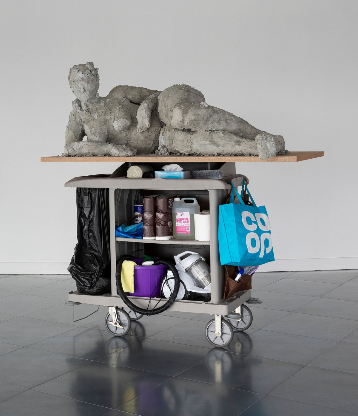 A female figure lying on her side on a board, beneath her a cleaning trolley on wheels containing various items.