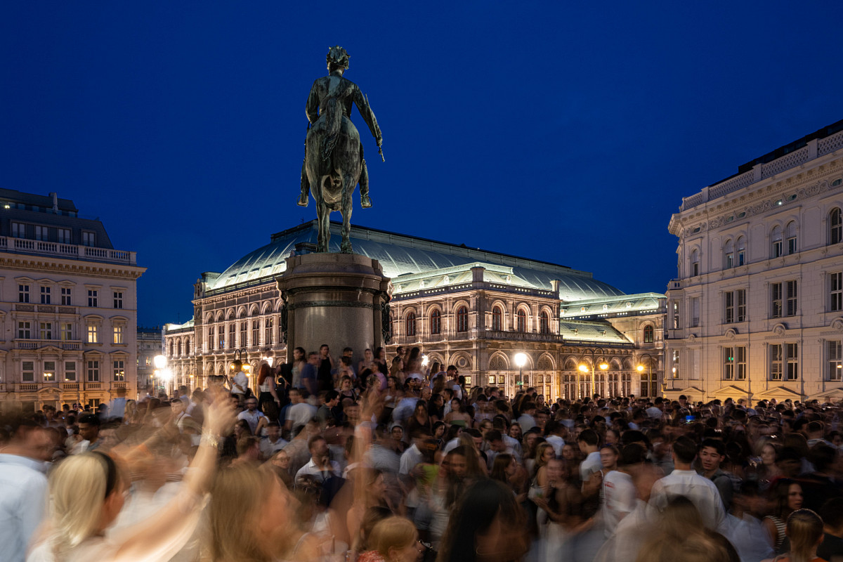 The ALBERTINA terrace in the evening, with a view of the illuminated Opera House and crowds of people dancing below the equestrian statue.