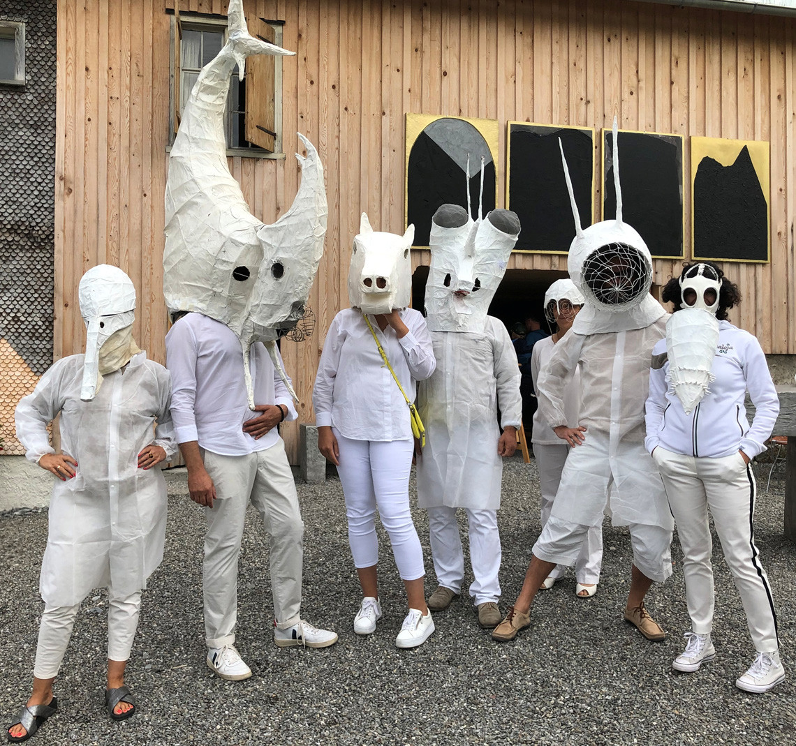 Verkleidete Menschen in weißen Gewändern mit weißen Masken vor einem Holzhaus.
