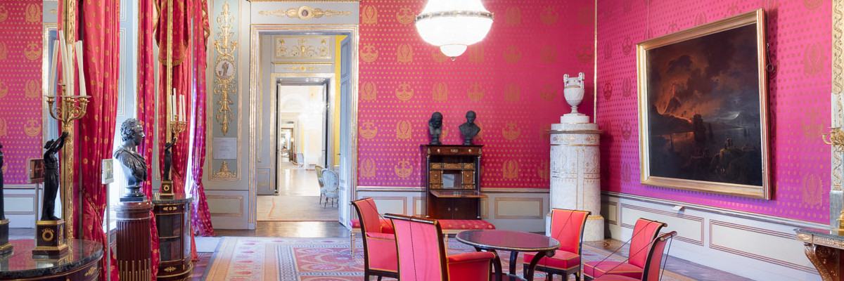 The audience room in the state rooms of the ALBERTINA is decorated entirely in red: The seating, walls and curtains are covered in red fabric. A large, dark painting with a golden border hangs on the wall. On two of the walls, a mirror covers almost the entire height of the wall. In front of the mirror on the floor are tables with candlesticks on them. White, gathered curtains and red, floor-length side panels with golden embroidery hang in front of the windows. Two large crystal chandeliers hang from the ceiling.